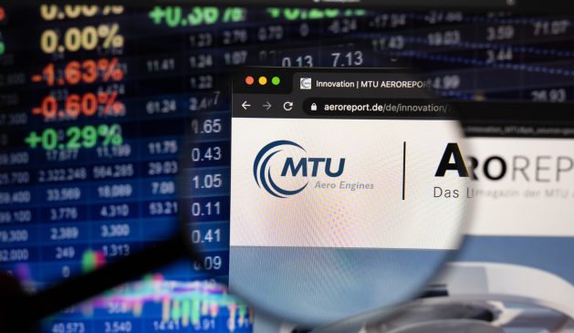 Firmenlogo der MTU Aero Engines AG auf einer Website mit verschwommenen Börsenentwicklungen im Hintergrund, gesehen auf einem Computerbildschirm durch ein Vergrößerungsglas