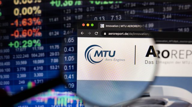 Firmenlogo der MTU Aero Engines AG auf einer Website mit verschwommenen Börsenentwicklungen im Hintergrund, gesehen auf einem Computerbildschirm durch ein Vergrößerungsglas