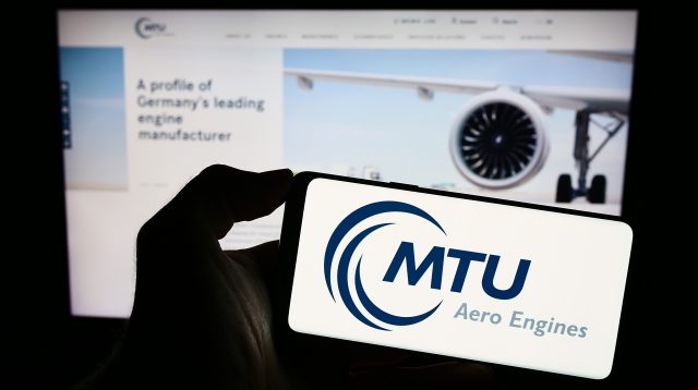 Eine Person hält ein Smartphone mit dem Logo des deutschen Luftfahrtunternehmens MTU Aero Engines AG auf dem Bildschirm vor einer Website