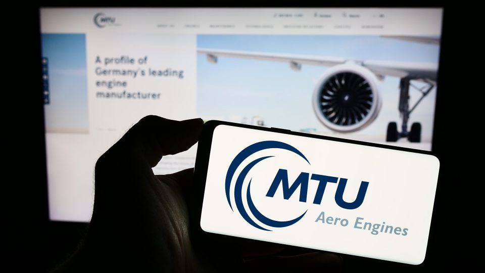 Eine Person hält ein Smartphone mit dem Logo des deutschen Luftfahrtunternehmens MTU Aero Engines AG auf dem Bildschirm vor einer Website