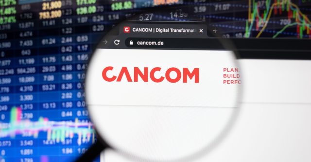 Cancom-Firmenlogo auf einer Website mit verschwommenen Börsenentwicklungen im Hintergrund, gesehen auf einem Computerbildschirm durch ein Vergrößerungsglas