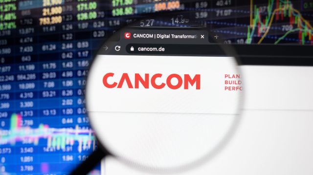 Cancom-Firmenlogo auf einer Website mit verschwommenen Börsenentwicklungen im Hintergrund, gesehen auf einem Computerbildschirm durch ein Vergrößerungsglas