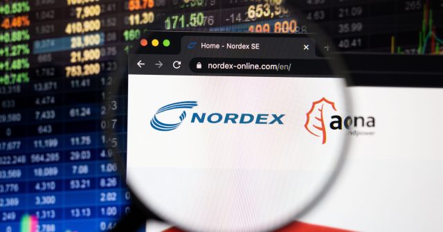 Nordex-Firmenlogo auf einer Website mit verschwommenen Börsenentwicklungen im Hintergrund, gesehen auf einem Computerbildschirm durch ein Vergrößerungsglas