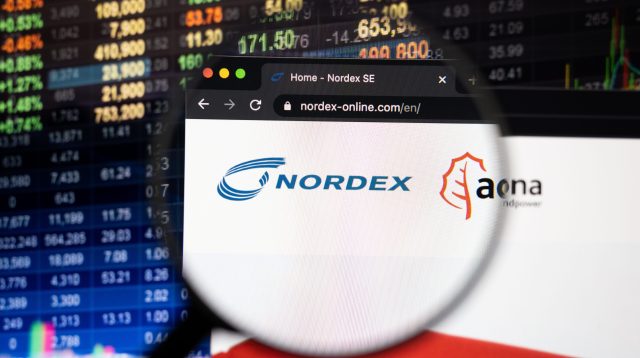 Nordex-Firmenlogo auf einer Website mit verschwommenen Börsenentwicklungen im Hintergrund, gesehen auf einem Computerbildschirm durch ein Vergrößerungsglas