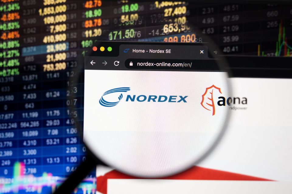 Nordex-Firmenlogo auf einer Website mit verschwommenen Börsenentwicklungen im Hintergrund, gesehen auf einem Computerbildschirm durch ein Vergrößerungsglas