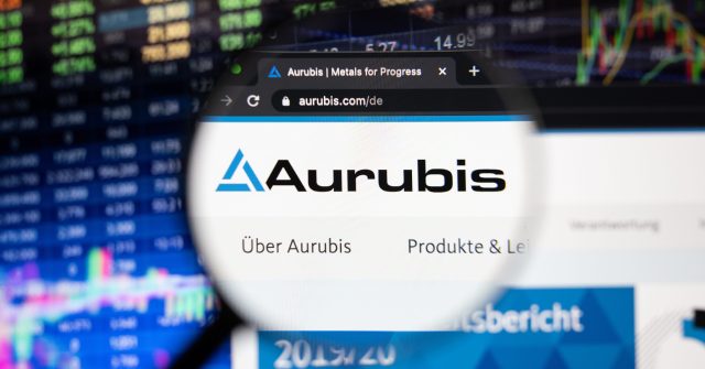 Aurubis-Firmenlogo auf einer Website mit verschwommenen Börsenentwicklungen im Hintergrund, gesehen auf einem Computerbildschirm durch ein Vergrößerungsglas