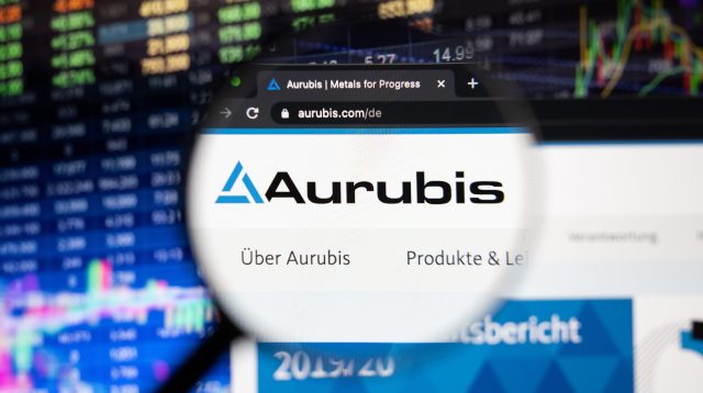 Aurubis-Firmenlogo auf einer Website mit verschwommenen Börsenentwicklungen im Hintergrund, gesehen auf einem Computerbildschirm durch ein Vergrößerungsglas