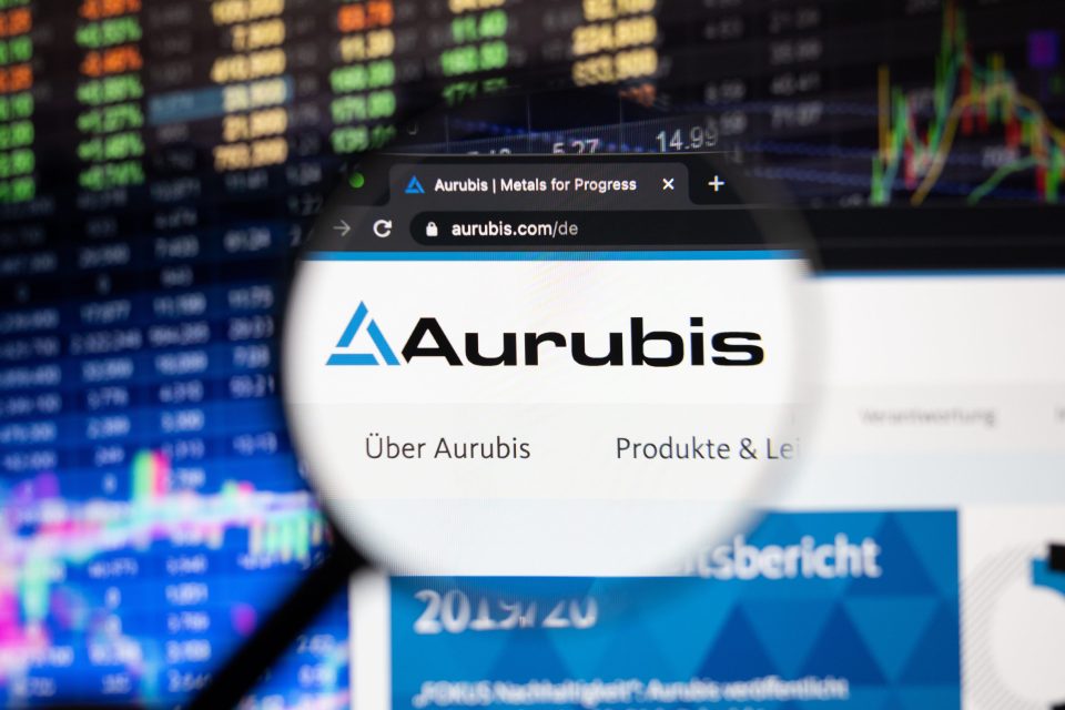 Aurubis-Firmenlogo auf einer Website mit verschwommenen Börsenentwicklungen im Hintergrund, gesehen auf einem Computerbildschirm durch ein Vergrößerungsglas