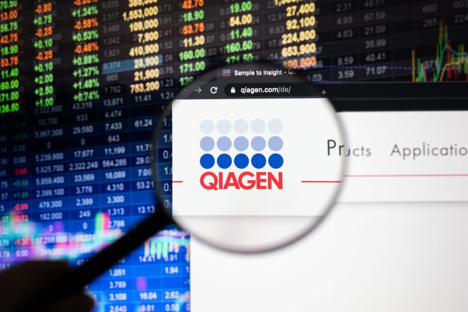 Das Qiagen-Firmenlogo auf einer Website mit verschwommenen Börsenentwicklungen im Hintergrund, gesehen auf einem Computerbildschirm durch ein Vergrößerungsglas