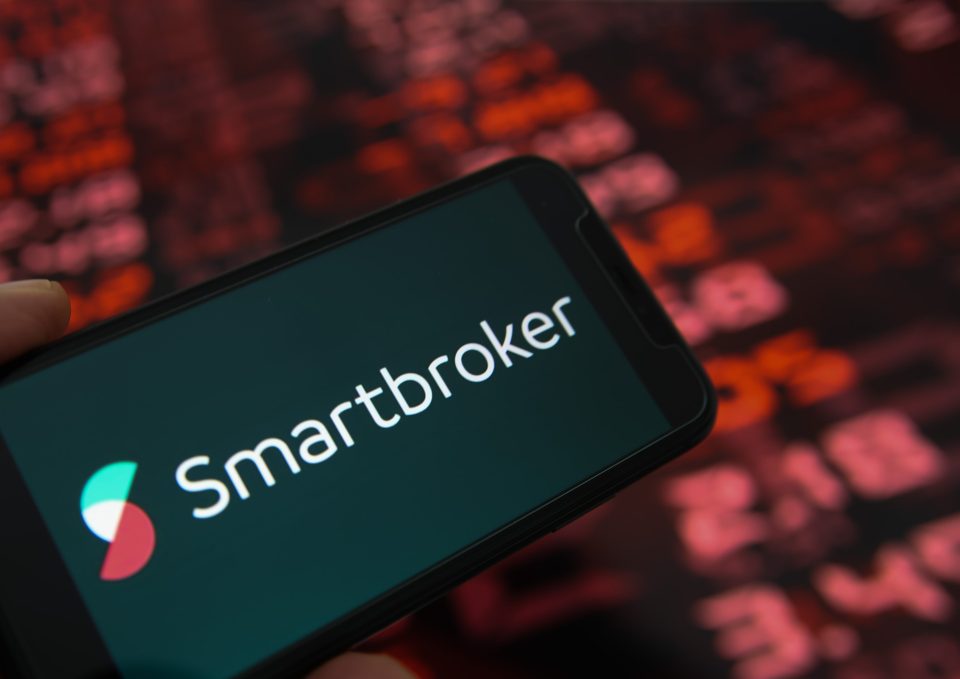 Nahaufnahme des Handy-Displays mit Logo-Schriftzug von Smartbroker, Börsenchart im Hintergrund