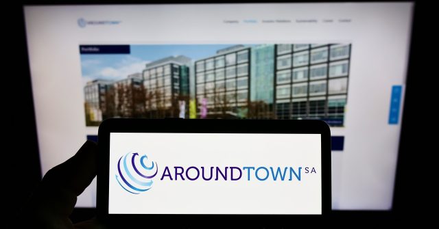 Eine Person hält ein Mobiltelefon mit dem Logo des Immobilienunternehmens Aroundtown SA auf dem Bildschirm vor einer Geschäftswebseite mit Fokus auf das Telefondisplay