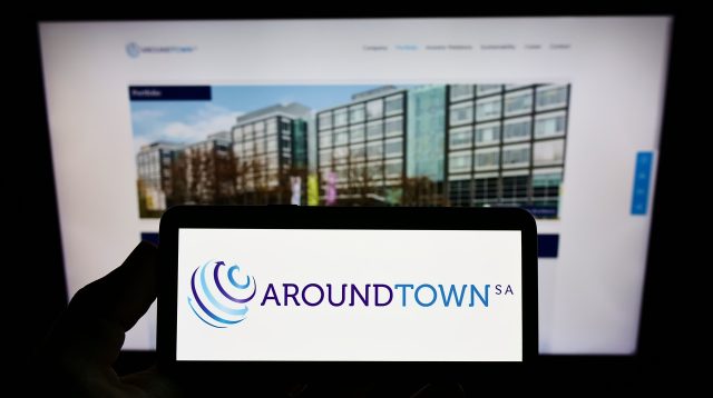 Eine Person hält ein Mobiltelefon mit dem Logo des Immobilienunternehmens Aroundtown SA auf dem Bildschirm vor einer Geschäftswebseite mit Fokus auf das Telefondisplay