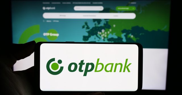 Eine Person hält ein Mobiltelefon mit dem Logo des ungarischen Finanzdienstleisters OTP Bank Group vor einer Webseite des Unternehmens mit Fokus auf das Telefondisplay