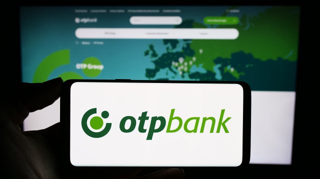 Eine Person hält ein Mobiltelefon mit dem Logo des ungarischen Finanzdienstleisters OTP Bank Group vor einer Webseite des Unternehmens mit Fokus auf das Telefondisplay