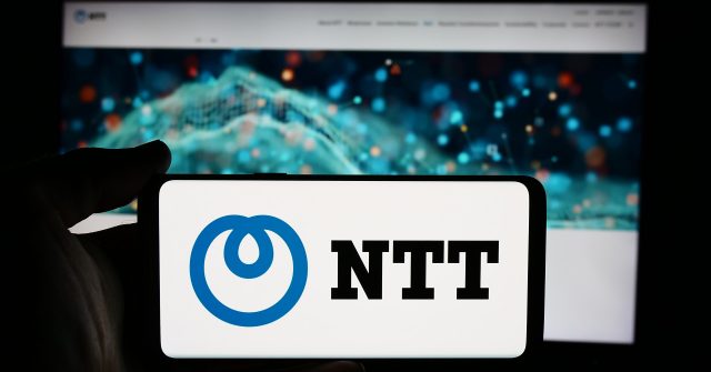 Eine Person hält ein Mobiltelefon mit dem Logo der Firma Nippon Telegraph and Telephone Corporation (NTT) vor einer Webseite mit Fokus auf das Telefondisplay