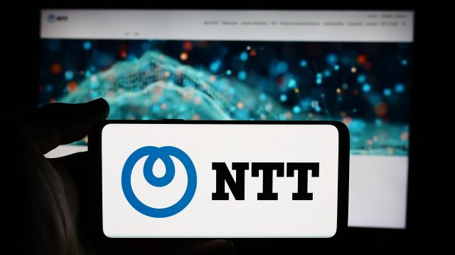 Eine Person hält ein Mobiltelefon mit dem Logo der Firma Nippon Telegraph and Telephone Corporation (NTT) vor einer Webseite mit Fokus auf das Telefondisplay