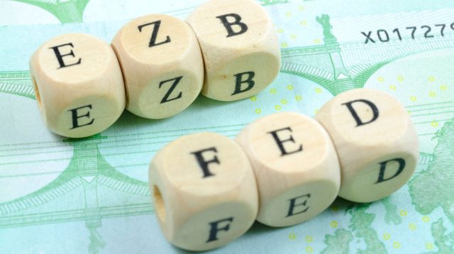 Die Buchstaben EZB und FED abgebildet auf Würfeln, welche auf einem 100€-Schein liegen